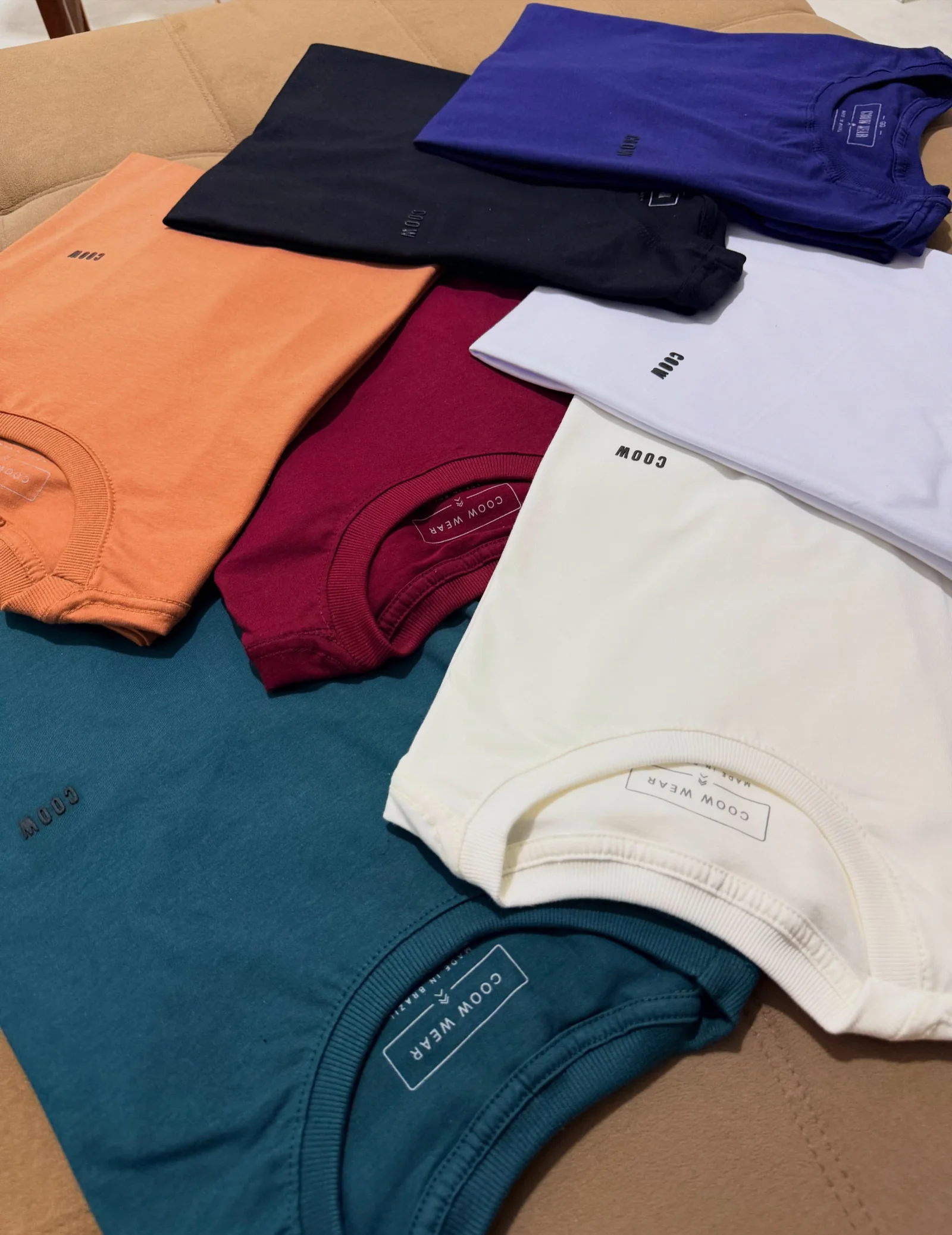 Peças reais dobradas da COOW WEAR exibindo gola canelada, bordado minimalista e variedade de cores